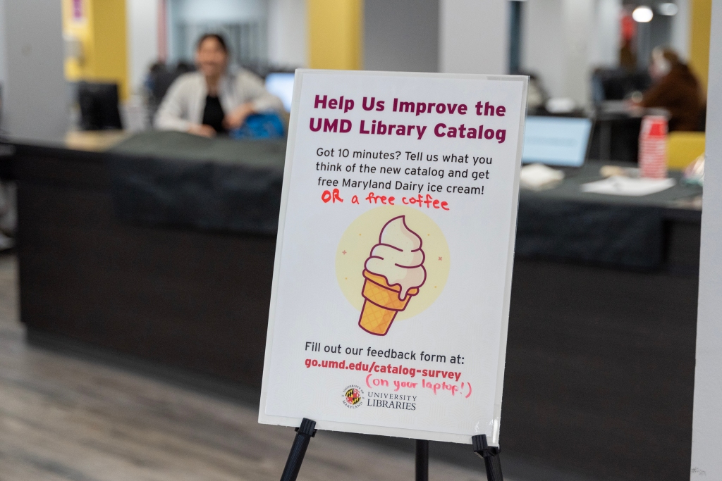 A sign on an easel advertises "Help us improve the UMD Library Catalog" with an image of an ice cream cone. The desk where user tests are conducted is in the background. 